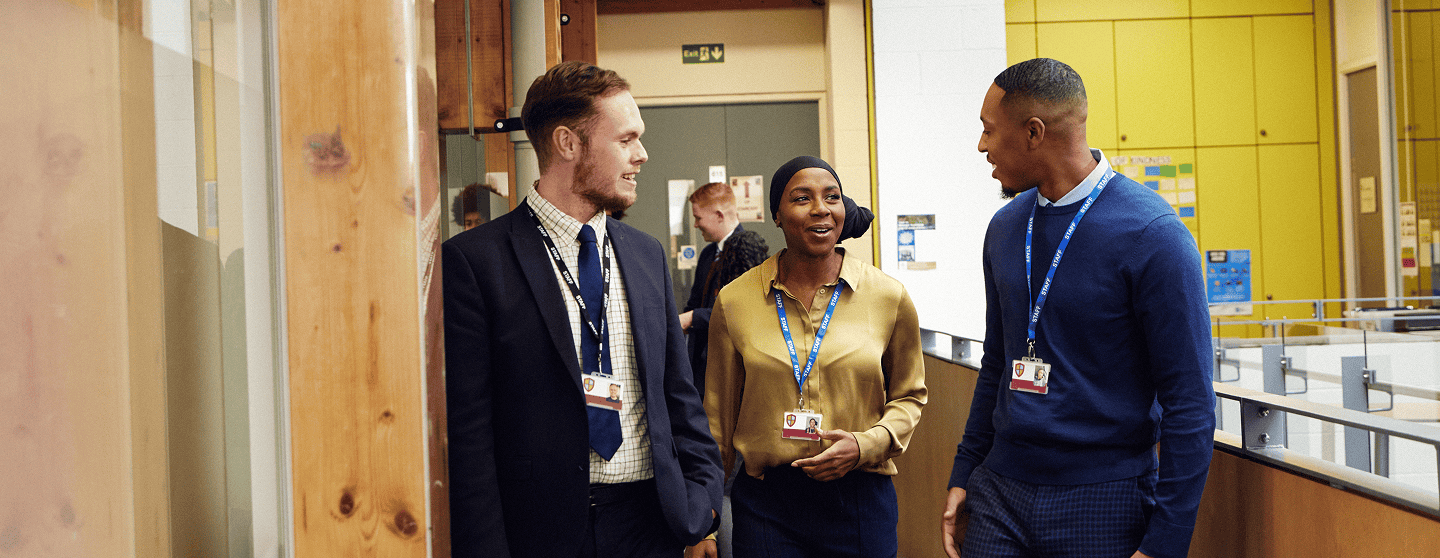 Three teachers walk together and talk in a school corridor, smiling and engaged in conversation.
